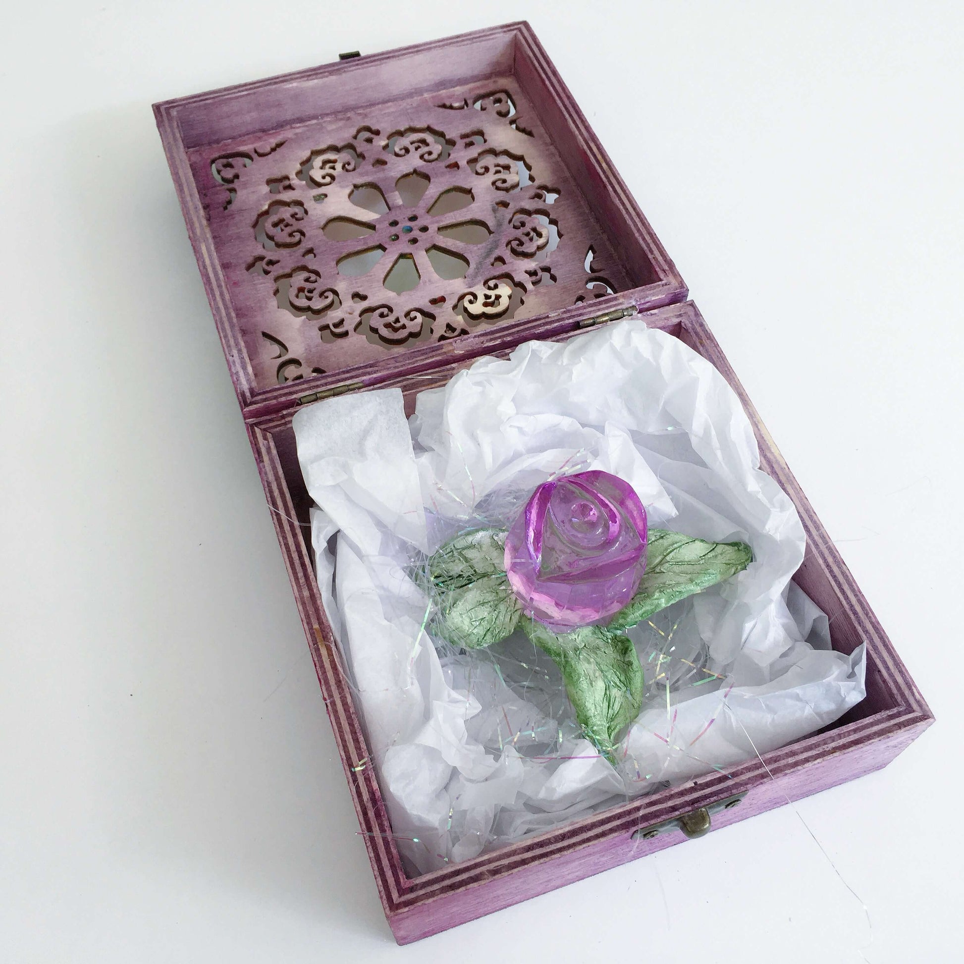 Contents of box, a purple glass rosebud with handmade ceramic leaves nestled in tissue paper and tinsel.