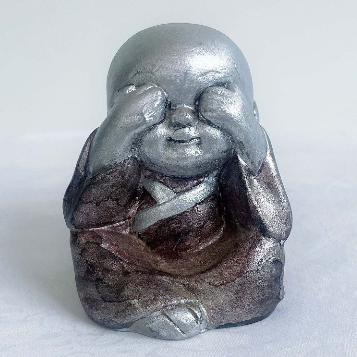 View of single monk, see no evil, painted silver figure with bronze robes, and hands covering the eyes.
