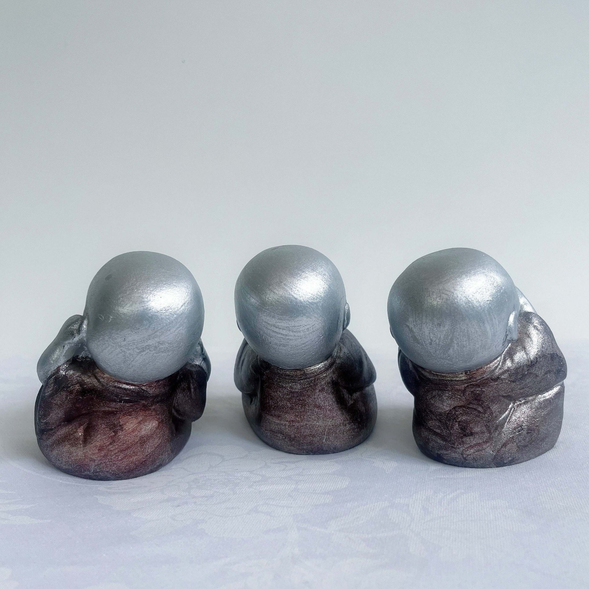 Back view of monk trio, painted silver figures with bronze robes, all sitting in a row.