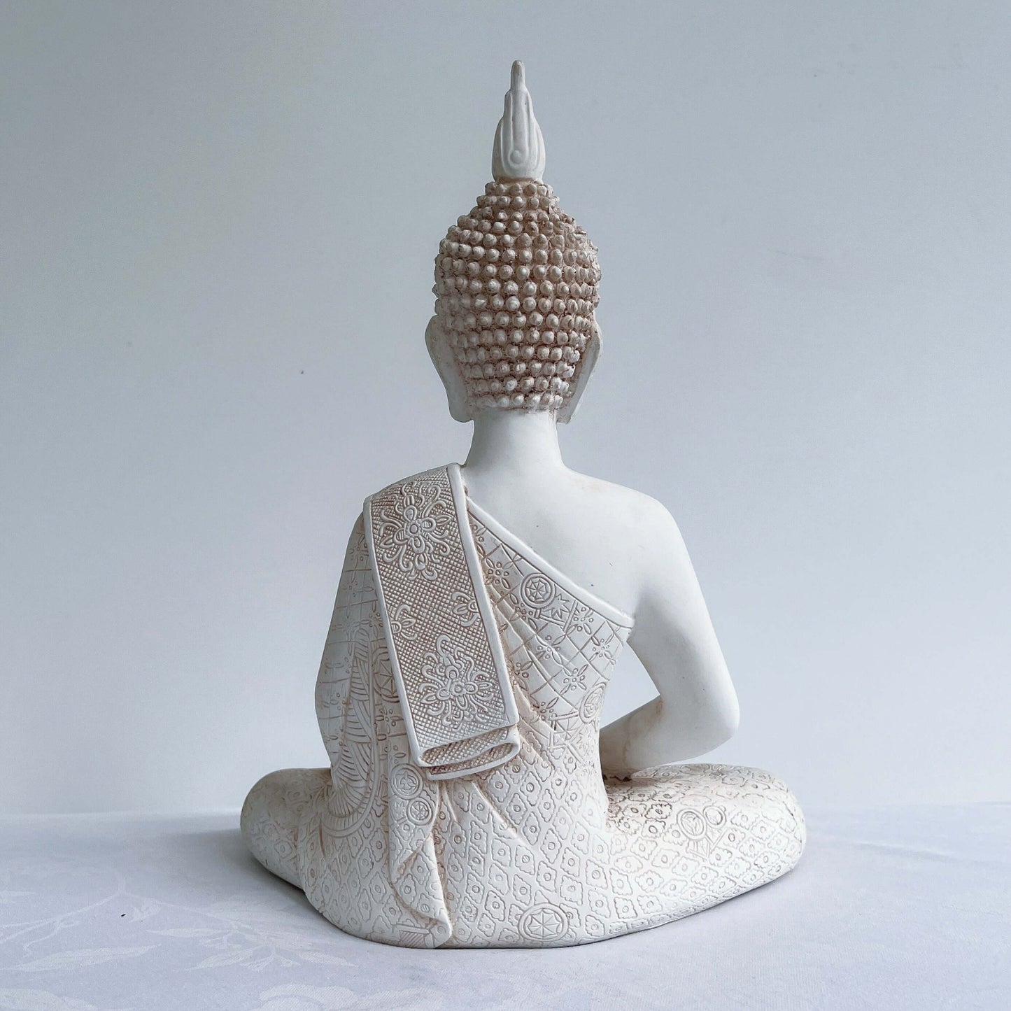 Back view of buddha in lotus position, natural off white coloring, ornate robes, with ushnisha on head.