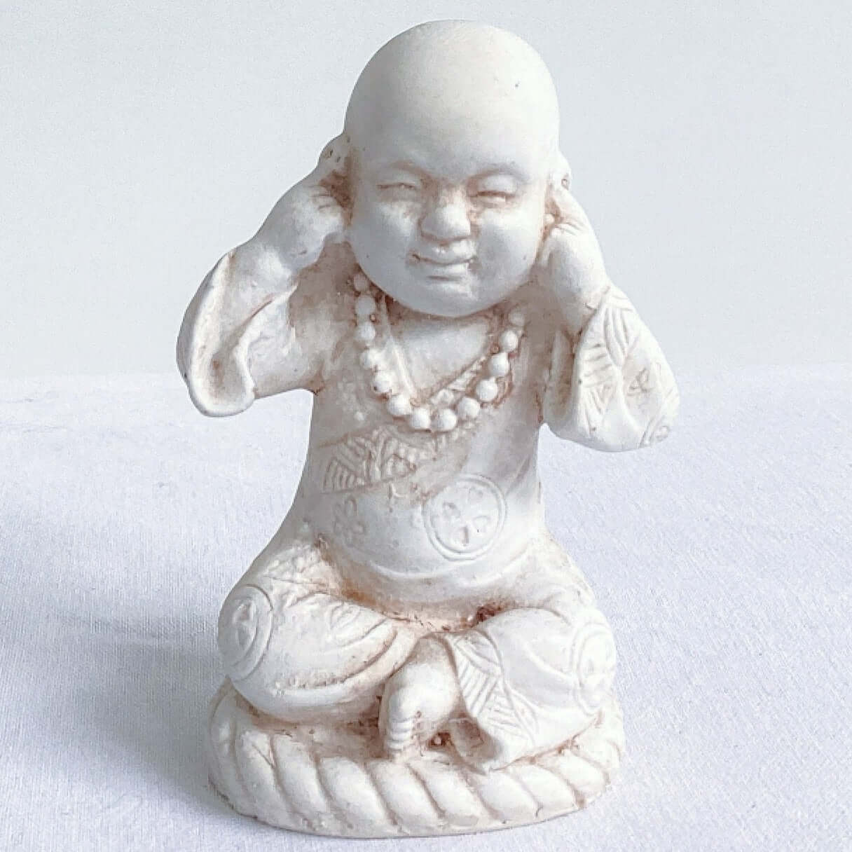 View of single monk, hear no evil, natural off white colors, with ornate robes, and hands covering the ears.
