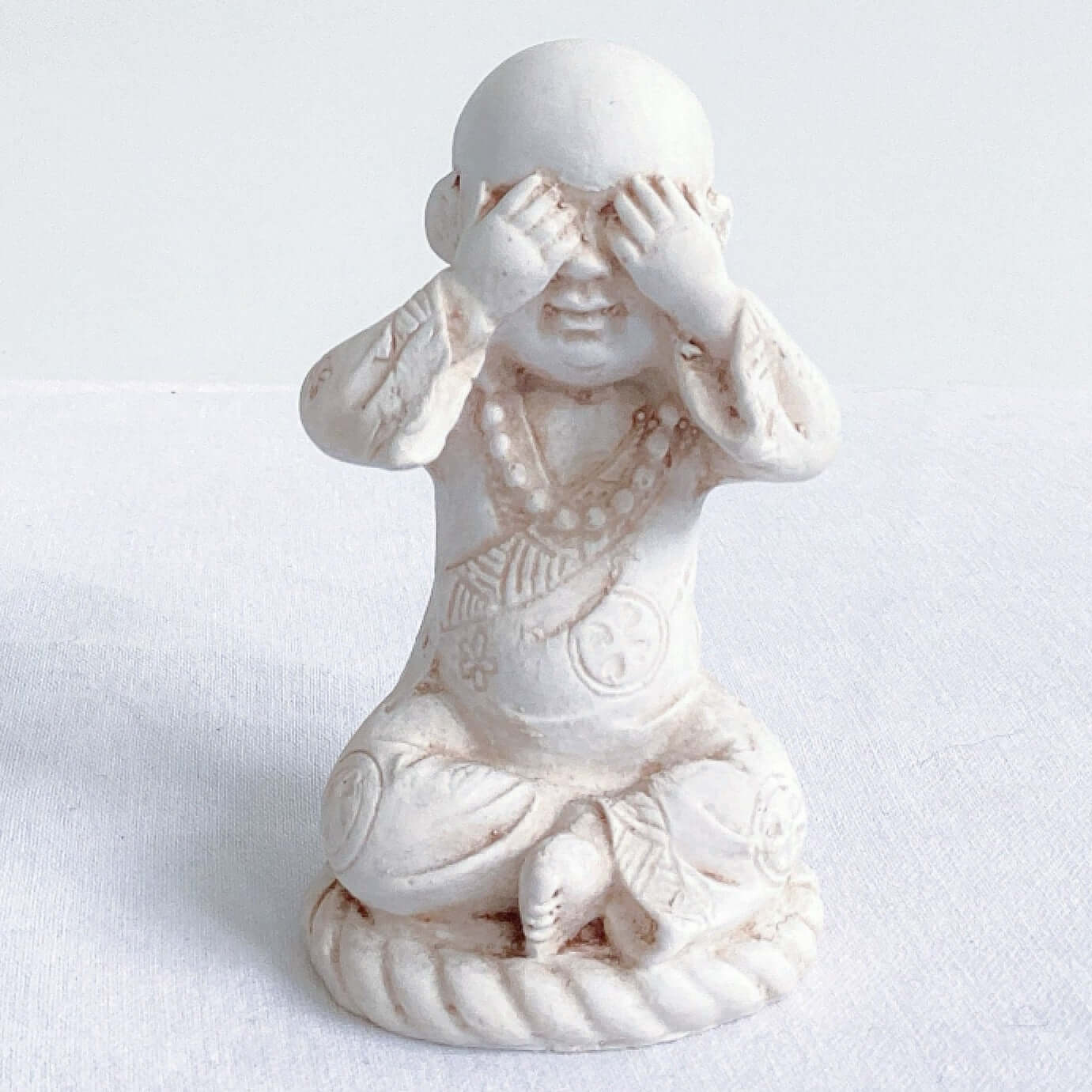 View of single monk, see no evil, natural off white colors, with ornate robes, and hands covering the eyes.