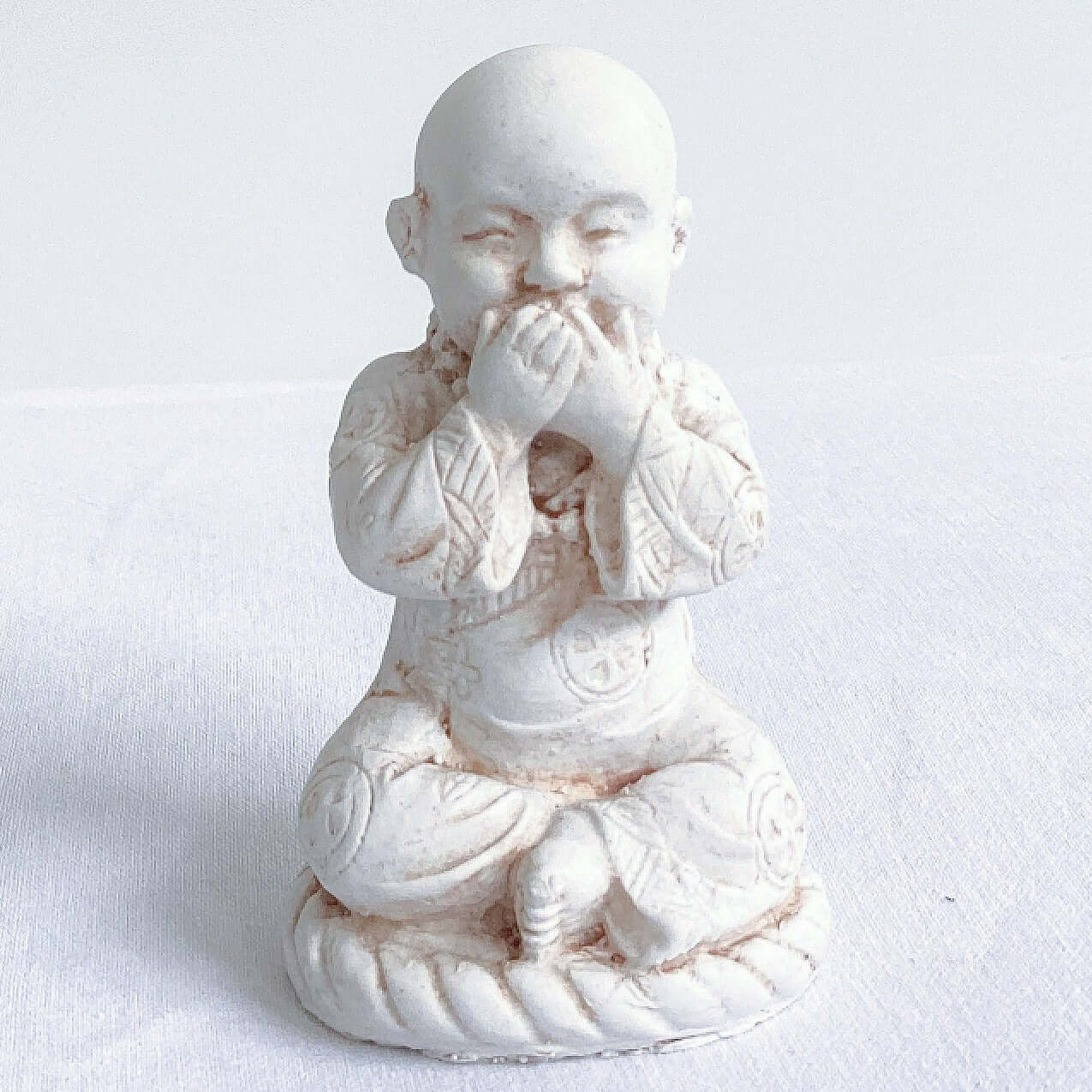 View of single monk, speak no evil, natural off white colors, with ornate robes, and hands covering the mouth.