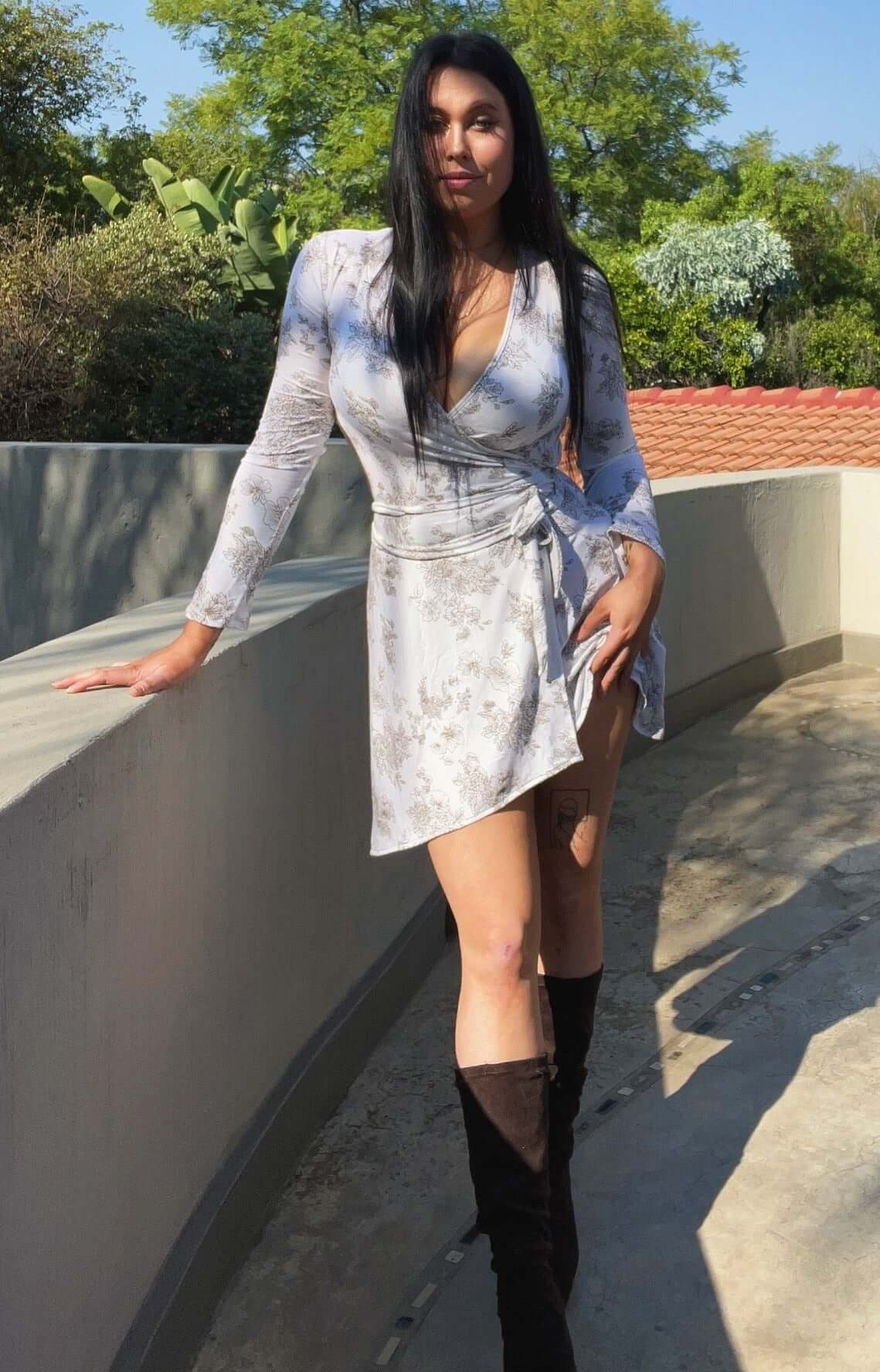 Front view of wrap dress, model standing and wearing long brown boots, trees behind.