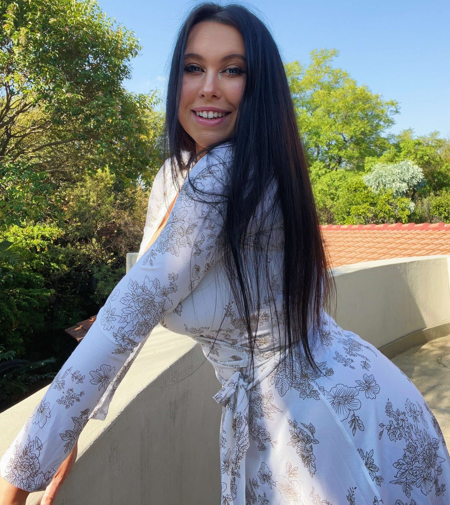 Side view of wrap dress, model leaning into a low wall, trees behind.