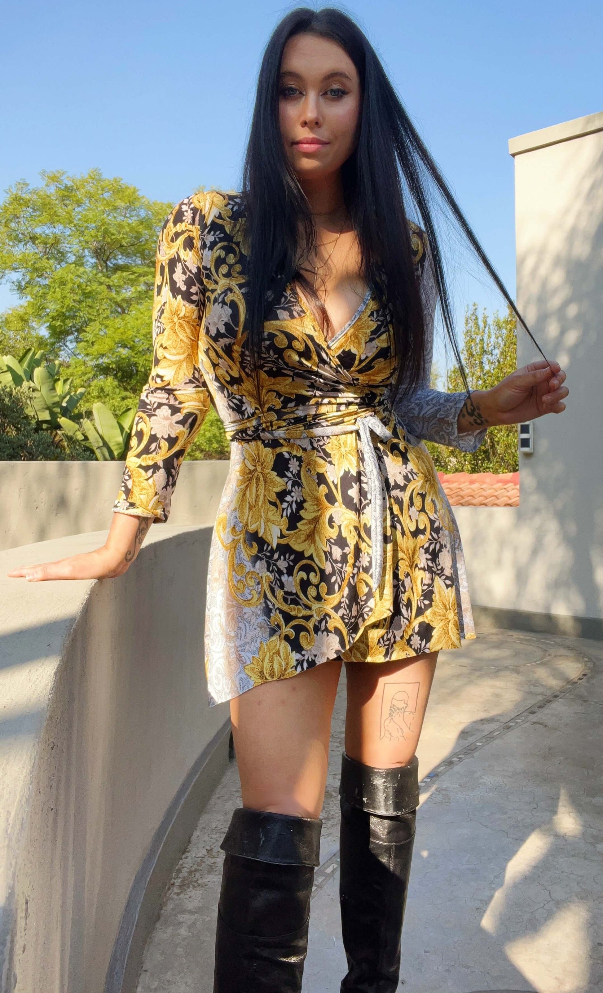 Front view of wrap dress with model wearing thigh-high boots, resting one hand on a low wall, trees in the background. 