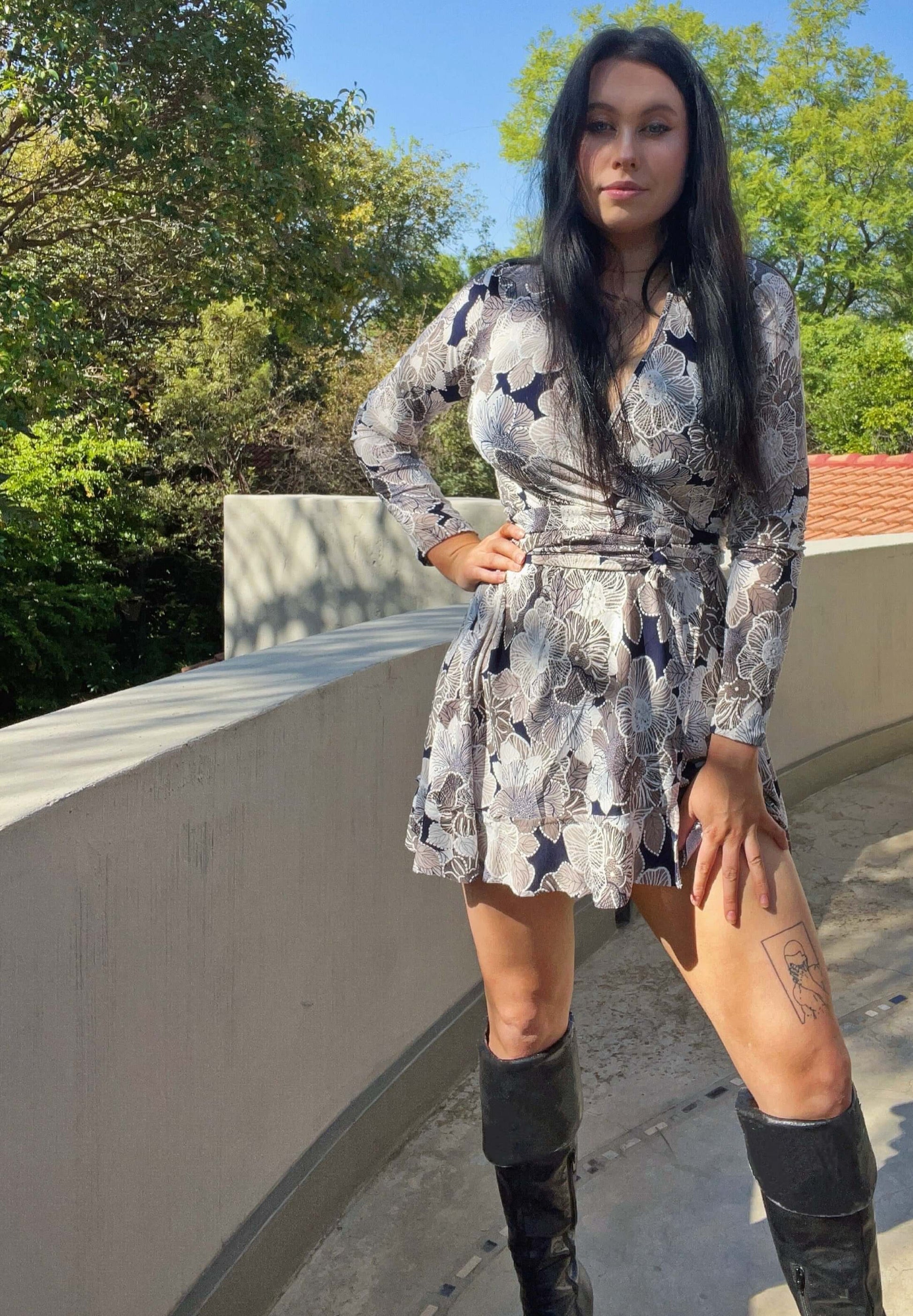 Front view of wrap dress, model wearing long thigh high boots, mid thigh length, trees and sky in the background.